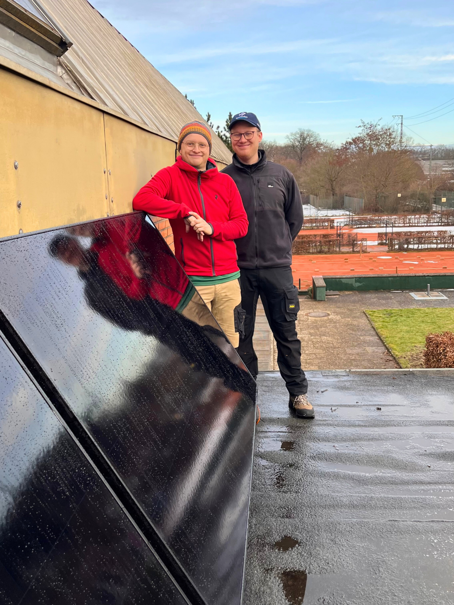 Zwei Personen, die lächelnd auf einem Flachdach neben den Panelen einer Balkon-Solaranlage stehen.