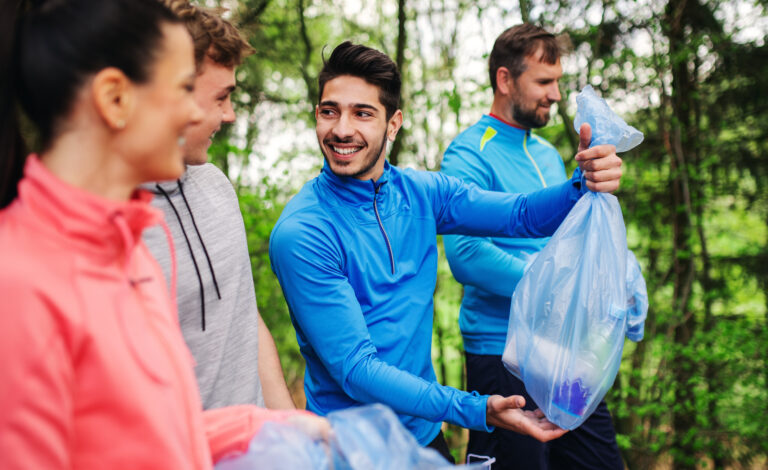 Eine Gruppe fröhlicher Sportler unterhält sich im Wald, während einer von ihnen einen blauen Plastikmüllsack mit gesammeltem Abfall hochhält.