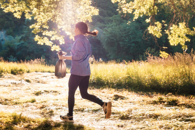 Eine joggende Frau läuft bei Sonnelicht mit einem gefüllten Müllsack in der Hand über eine sonnige Lichtung.