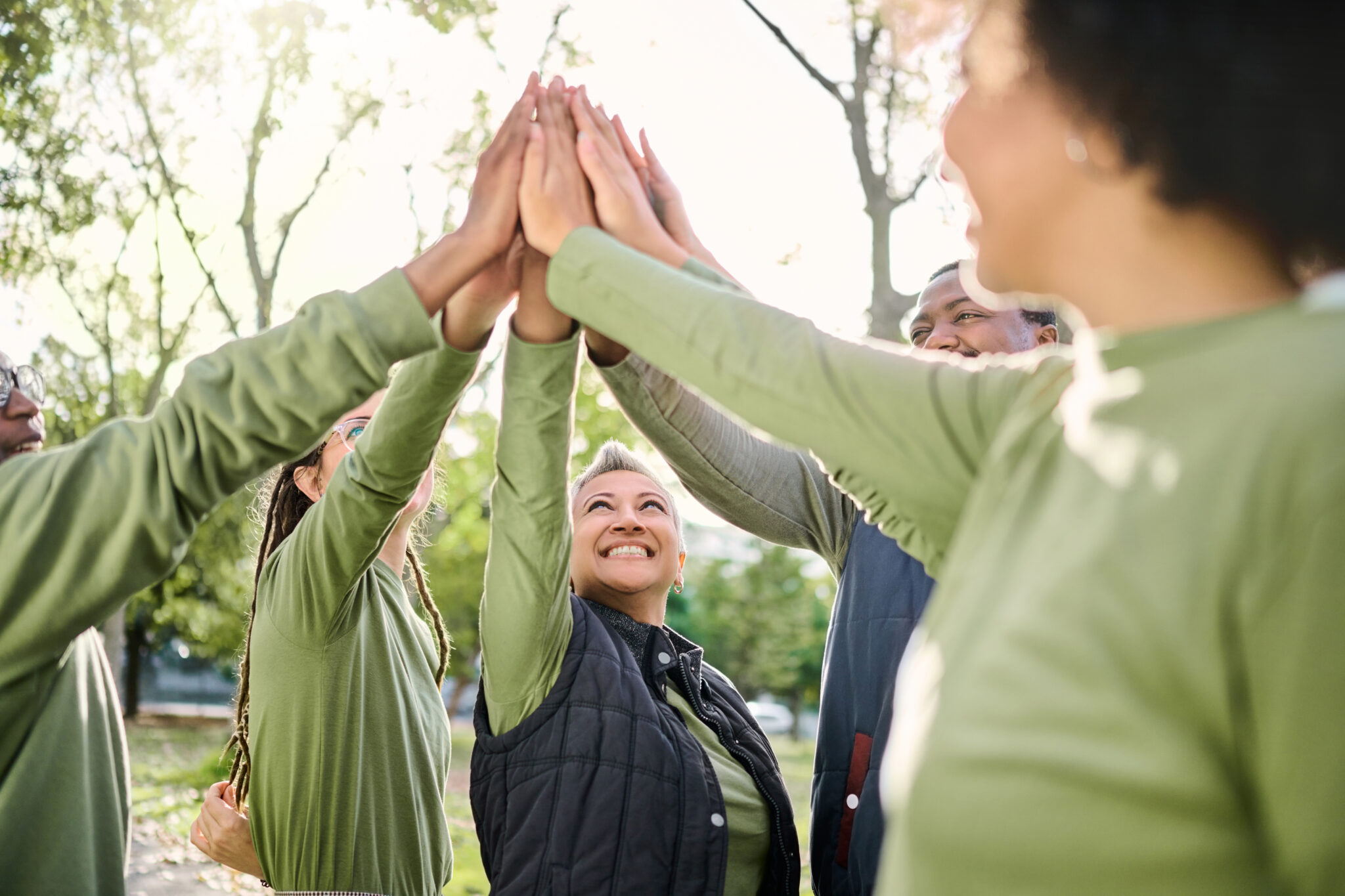 Eine Gruppe in passenden grünen Hemden, die im Kreis stehen und ihre Hände in die Mitte legen, um High-Fives zu geben, um Teamarbeit und Feierlichkeit zu zeigen, während sie in einem Park im Freien sind.