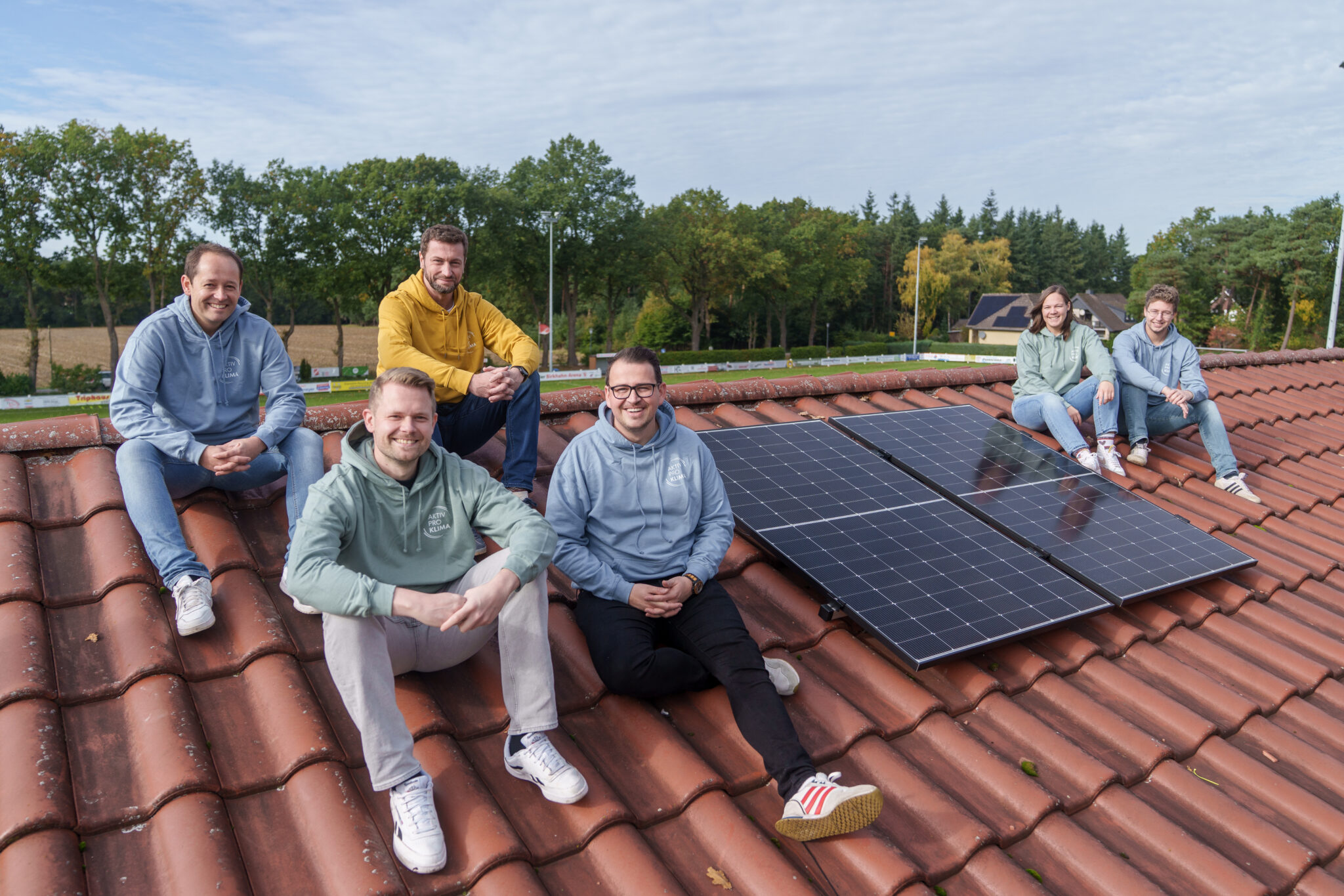 Gruppe von sechs Engagierten eines Sportvereins in bunten Hoodies sitzt auf dem Dach des Vereinsheims direkt neben zwei installierten Solarmodulen.