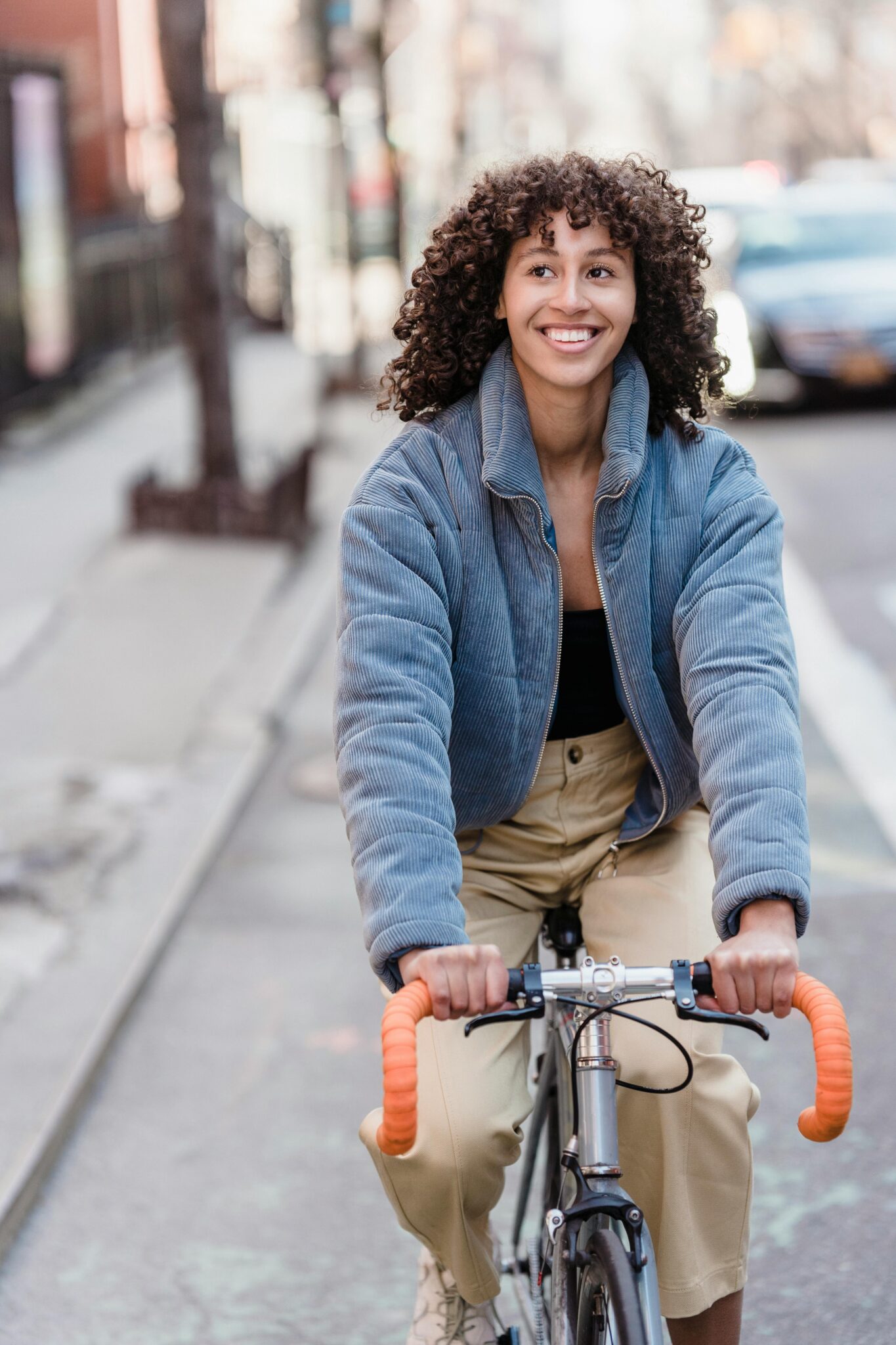 Eine Aufnahme einer jungen Frau mit lockigem Haar, die lächelt und Fahrrad fährt. Sie trägt eine blaue Cord-Steppjacke und beigefarbene Hosen.