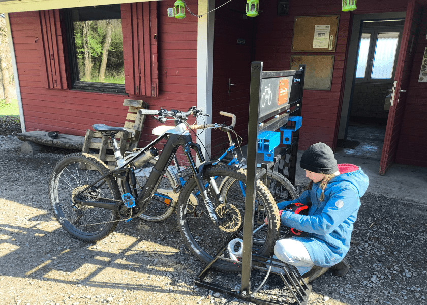 Ein Kind in einer blauen Jacke kniet vor einer Fahrradstation und hantiert am Vorderrad eines Mountainbikes vor einer roten Holzhütte.