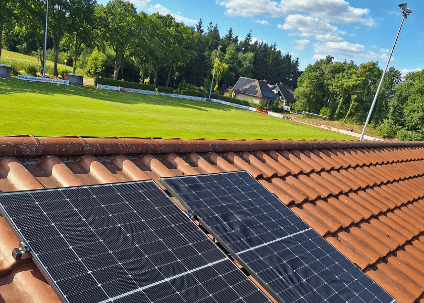 Die Solarmodule eines Balkonkraftwerks sind auf einem roten Ziegeldach montiert, im Hintergrund ist ein grüner Sportplatz zu sehen.