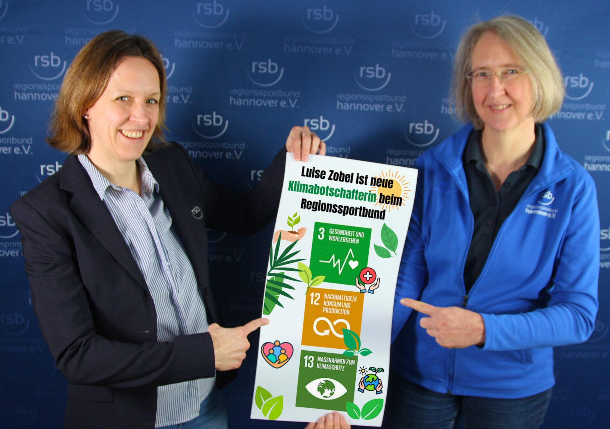 Zwei lächelnde Frauen halten ein Plakat mit den Klimazielen 3, 12 und 13 in der Hand. Sie zeigen mit der jeweils anderen Hand auf das Plakat.