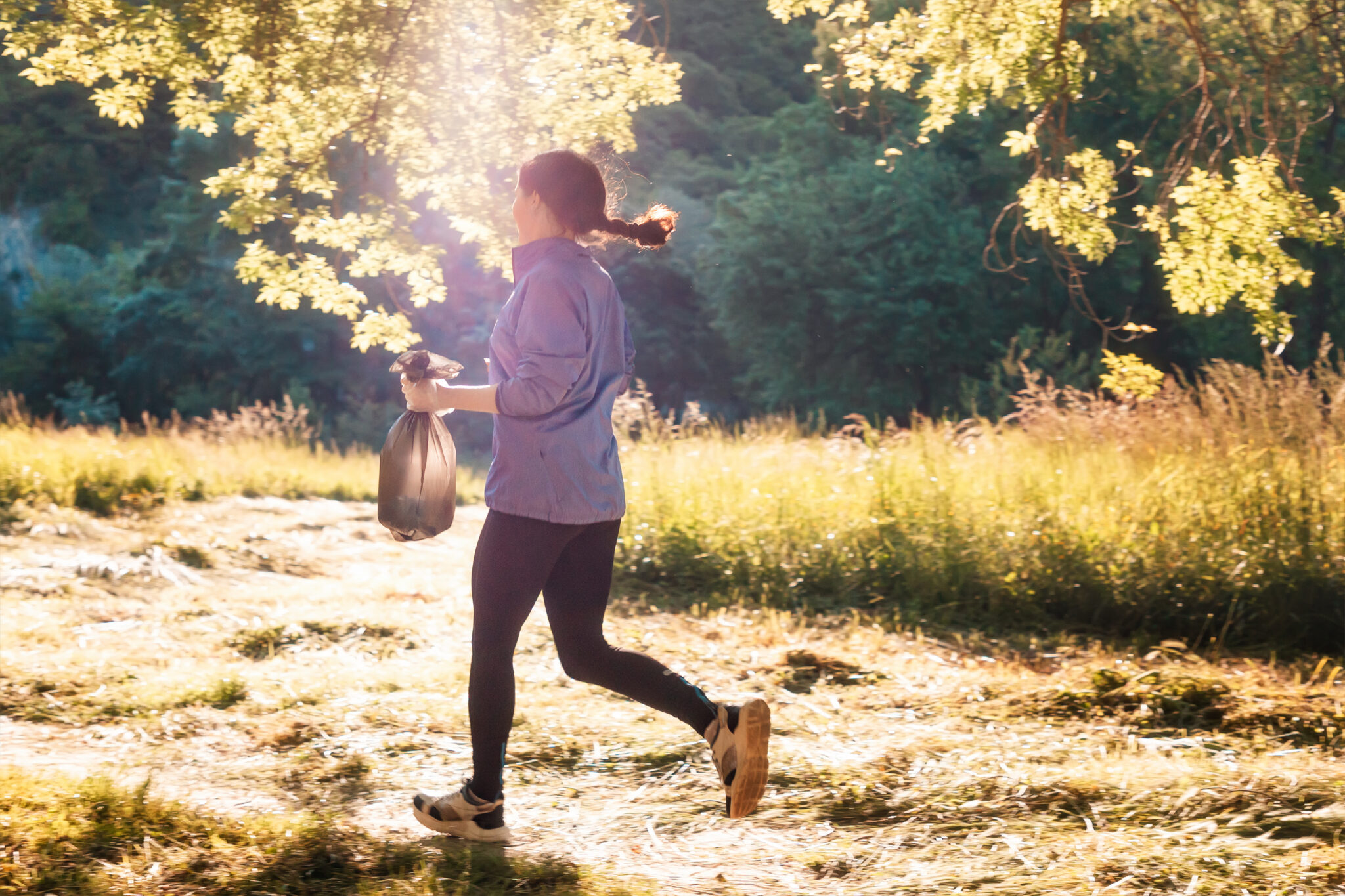 Eine joggende Frau (Ploggerin) läuft bei Sonnenaufgang mit einem gefüllten Müllsack in der Hand über eine sonnige Lichtung.