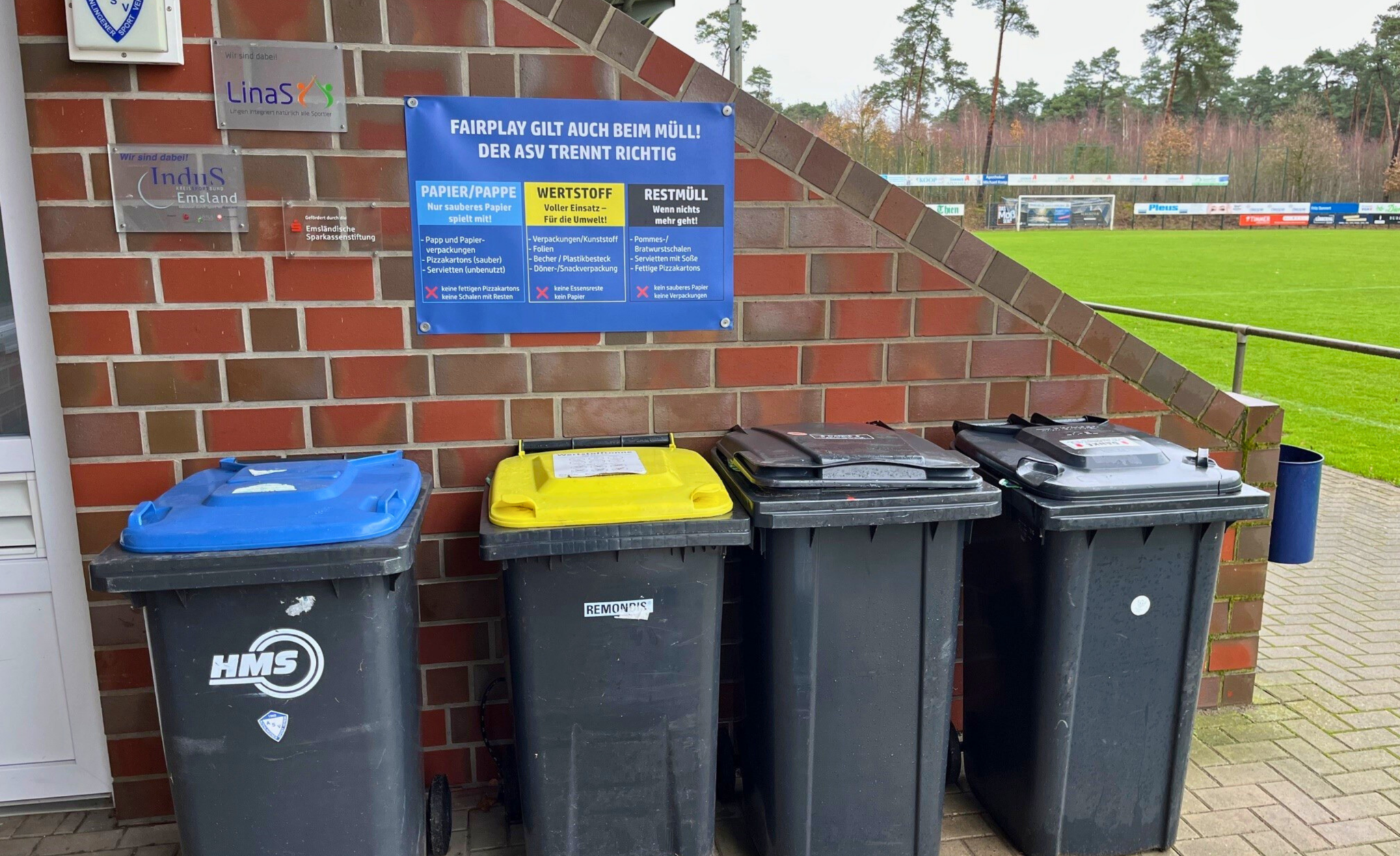 4 Mülltonnen vor einer gemauerten Wand an einem Sportplatz und ein Schild über den Tonnen zur korrekten Mülltrennung.