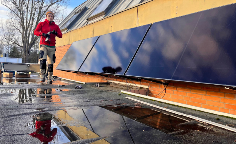 Eine Personen auf einem Flachdach mit vielen Pfützen. An der Wand dahinter sind Solarpanele befestigt.