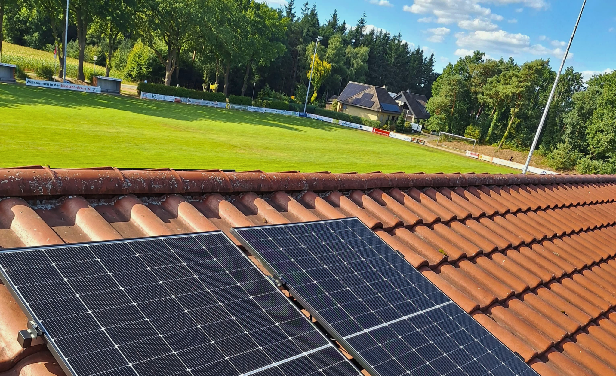Die Solarmodule eines Balkonkraftwerks sind auf einem roten Ziegeldach montiert, im Hintergrund ist ein grüner Sportplatz zu sehen.