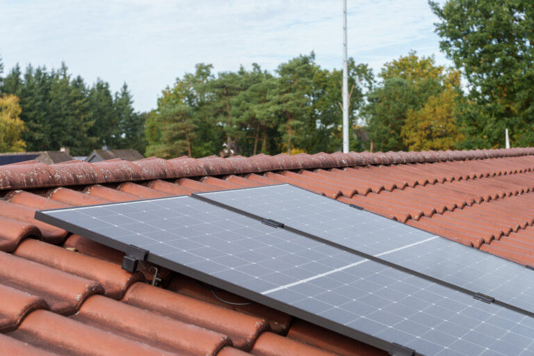 Zwei Solarmodule einer Balkkon-Solaranlage auf einem mit roten Ziegeln gedeckten Hausdach vor einer Kulisse aus grünen Bäumen.