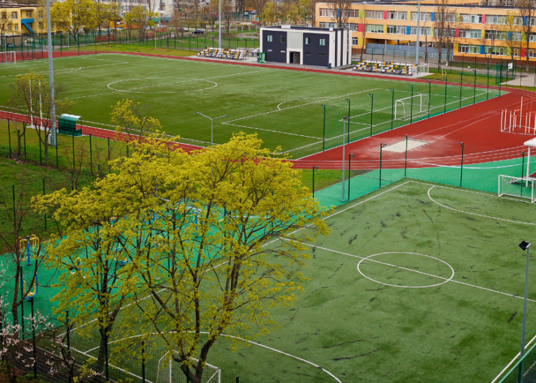 Eine Nahaufnahme einer modernen Sportanlage mit mehreren Spielfeldern, einer Laufbahn und einem farbenfrohen Schulgebäude im Hintergrund.