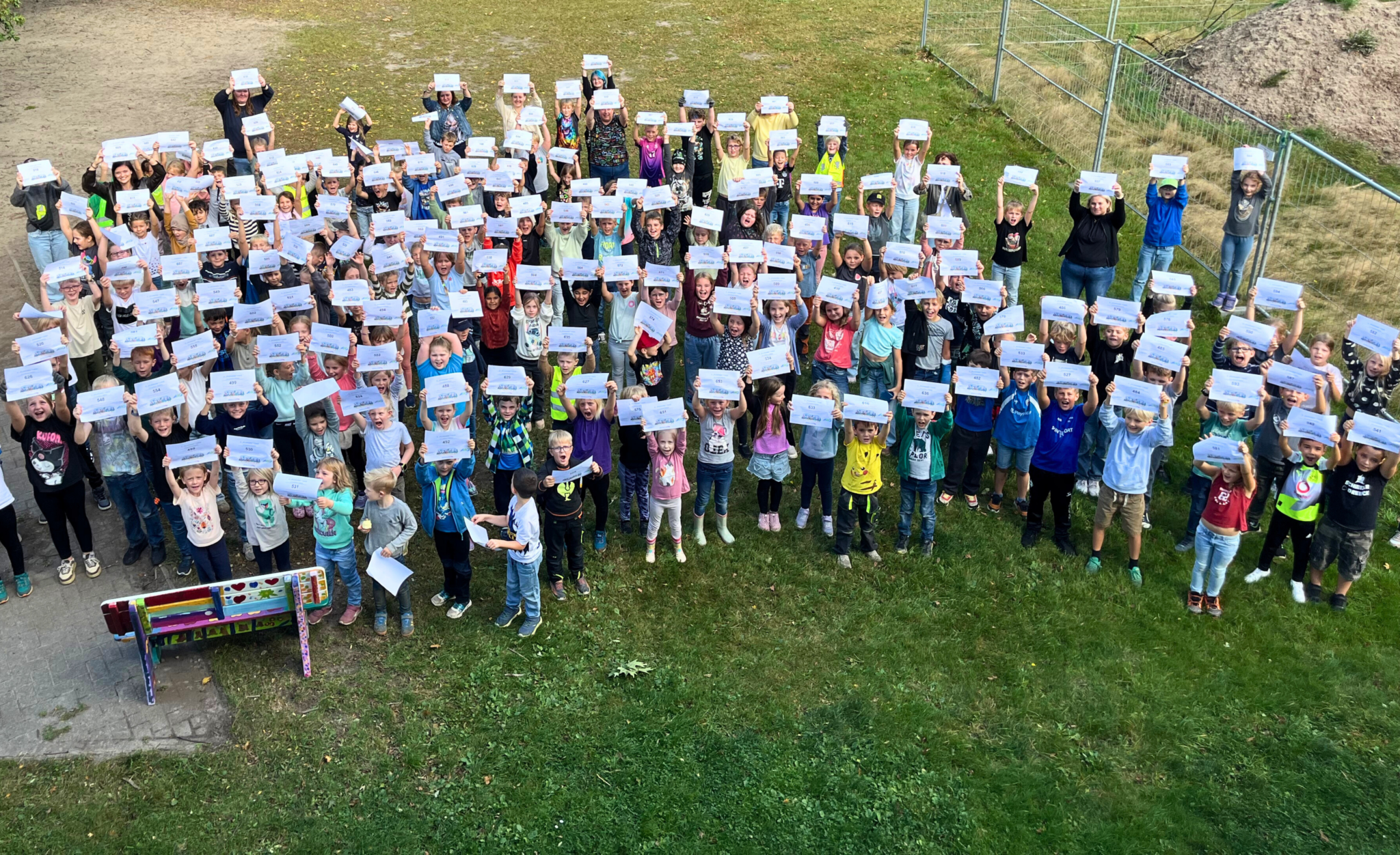Eine sehr große Gruppe von Schulkindern steht auf einer Wiese und hält gemeinsam weiße Urkunden in die Höhe.