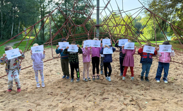 Kindergruppe auf einem sandigen Spielplatz vor einem großen, roten Seilklettergerüst. Alle Kinder stehen in einer Reihe und halten weiße Urkunden mit Nummern hoch, die ihre Gesichter komplett verdecken.