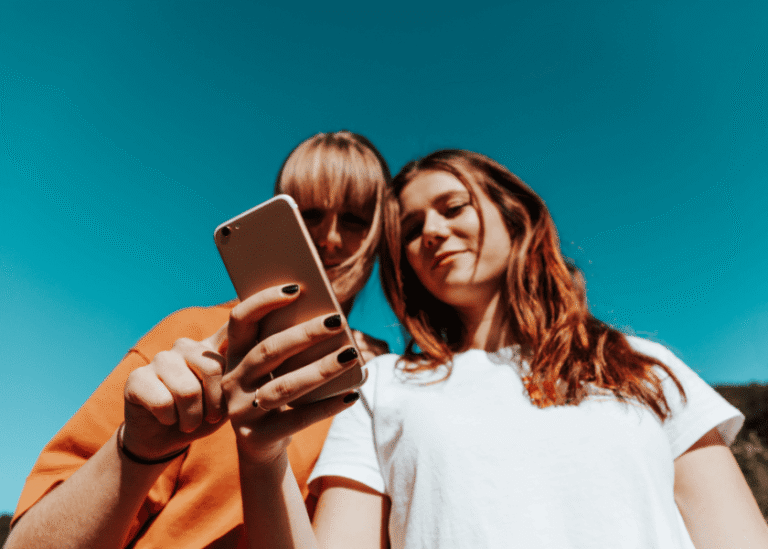 Zwei junge Frauen blicken von oben herab auf ein Smartphone vor einem strahlend blauen Himmel.