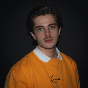 Nahaufnahme eines jungen Mannes mit dunklen Haaren, markantem Schnurrbart und Ohrringen. Er trägt einen gelb-orangefarbenen Pullover mit weißem Kragen und blickt direkt in die Kamera.