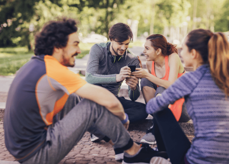 Eine Gruppe von vier jungen Erwachsenen in Sportkleidung sitzt im Freien zusammen und schaut lachend auf ein Smartphone.